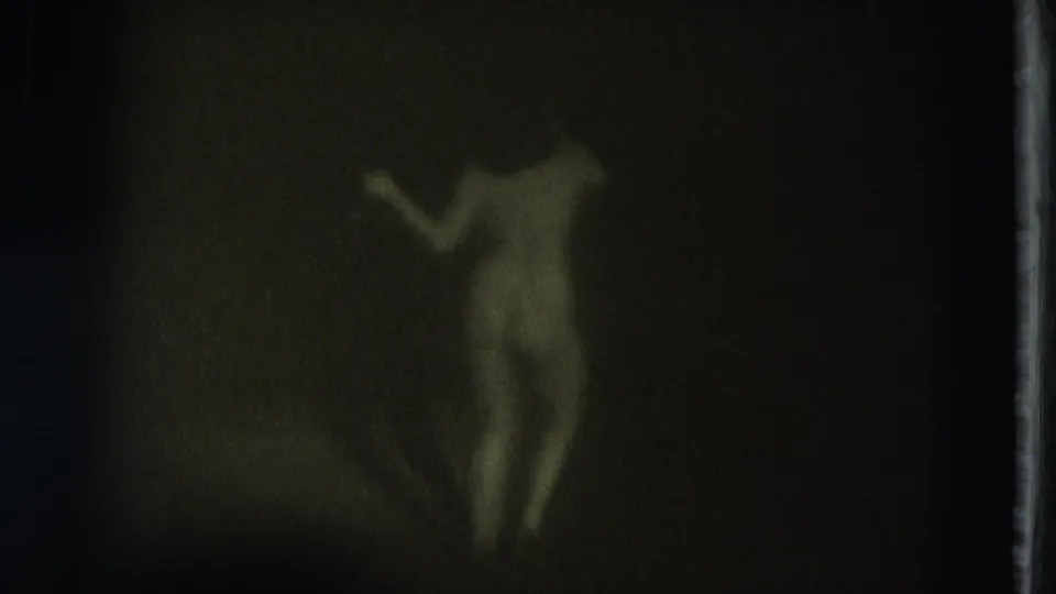 Green-tinted 1975 8mm frame from San Francisco showing a nude adult woman in soft focus with one arm raised against a dark interior wall.