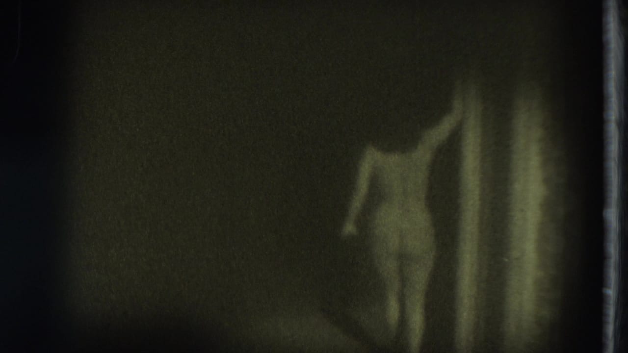 Green-toned 8mm frame shows a nude adult woman in a dark interior, her body partly silhouetted beside a vertical hallway or doorway edge.