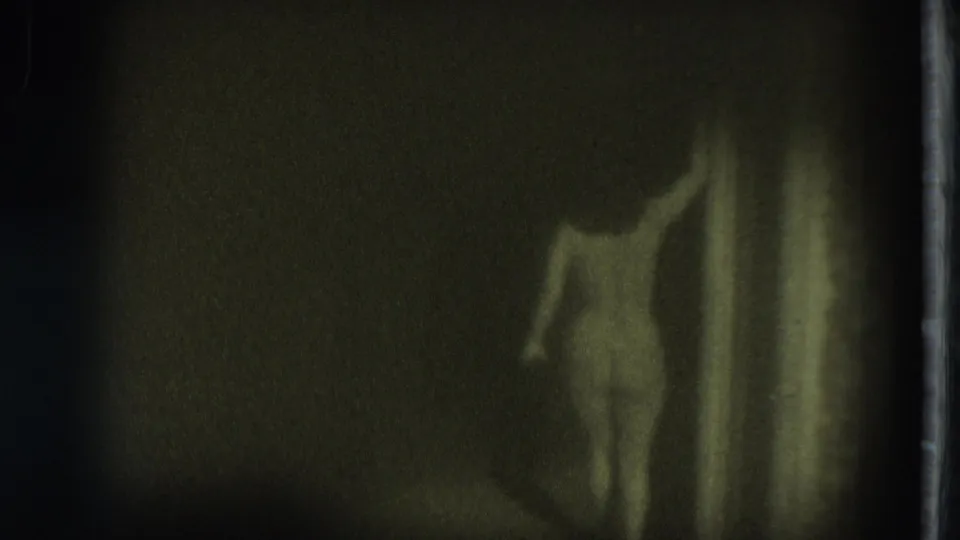 Green-toned 8mm frame shows a nude adult woman in a dark interior, her body partly silhouetted beside a vertical hallway or doorway edge.