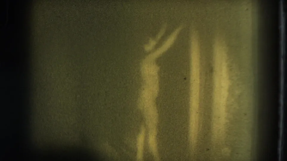 1975 8mm home-movie frame from San Francisco showing a nude adult woman dancing with arms raised in a yellow-green patch of light.