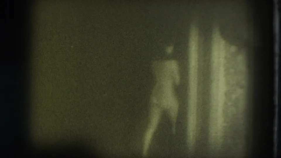 Grainy 8mm frame shows a nude adult woman moving away or turning in a dark San Francisco interior, with a bright vertical wall area beside her.