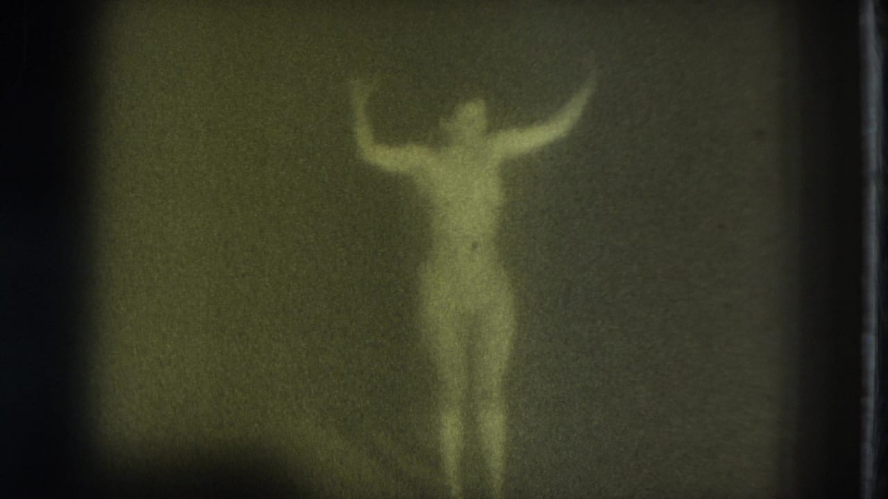 Blurred nude adult woman stands with arms raised in a dim green-tinted frame.