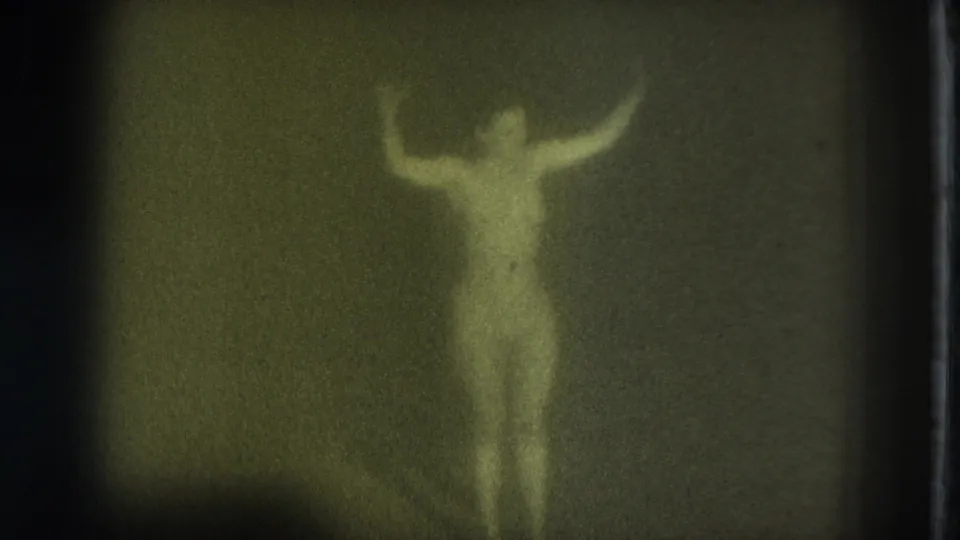 1975 San Francisco 8mm frame showing a blurred nude adult woman facing the camera with arms raised in a dark interior.