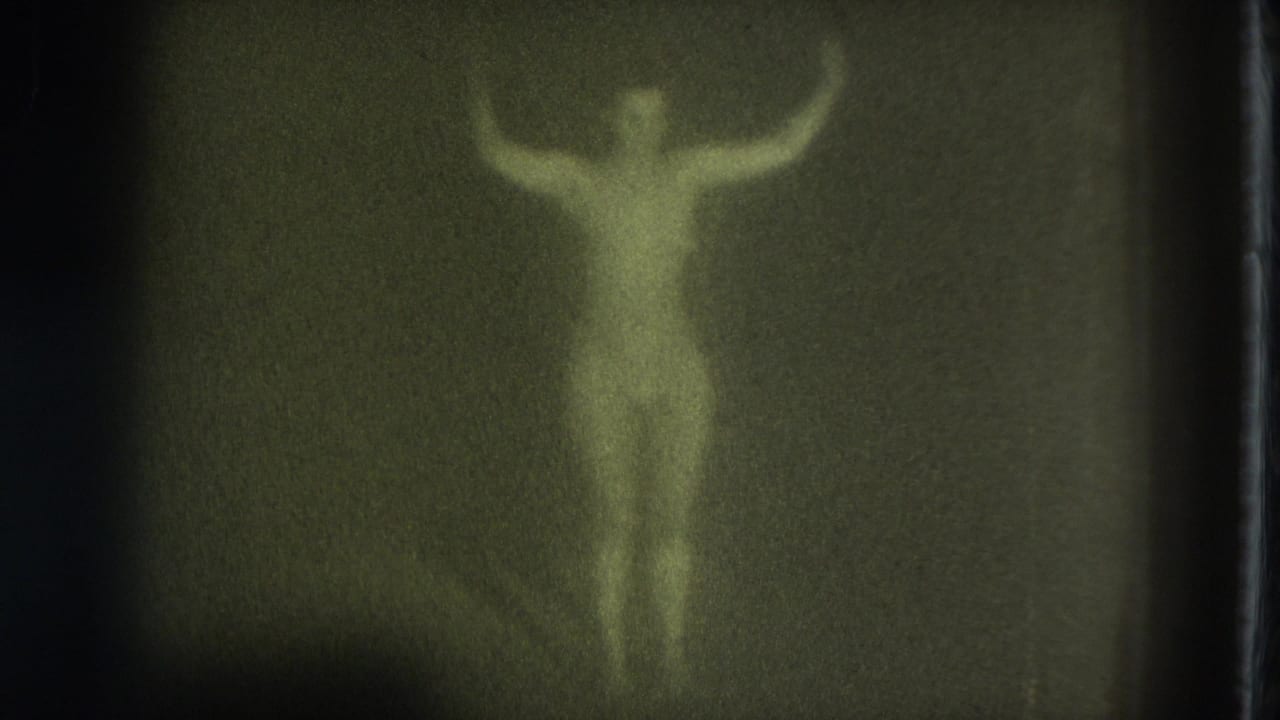 Green-tinted 1975 8mm frame of a nude adult woman standing in a dim room with arms lifted and details obscured by grain.
