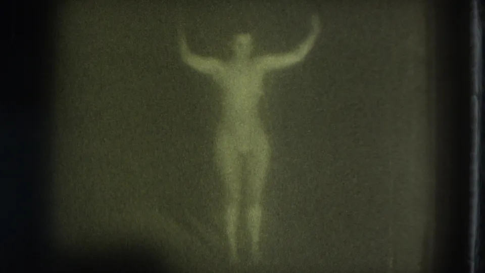 Green-tinted 1975 8mm frame of a nude adult woman standing in a dim room with arms lifted and details obscured by grain.