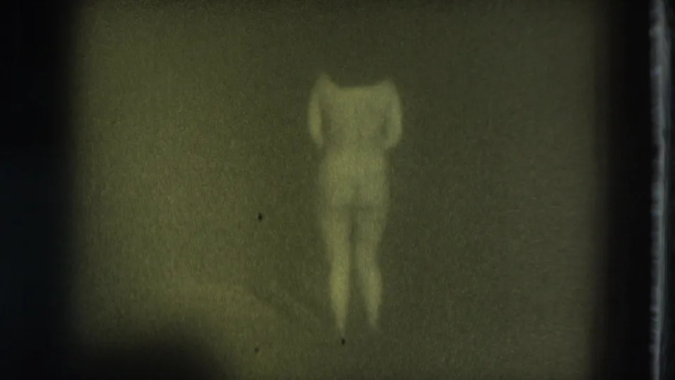 1975 San Francisco 8mm frame showing a nude adult woman in a dim interior, arms bent near her head and body softened by blur.