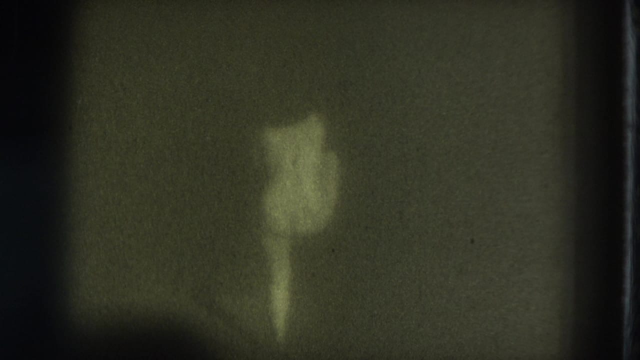 Distant nude adult woman standing on a dark stage under dim greenish light.
