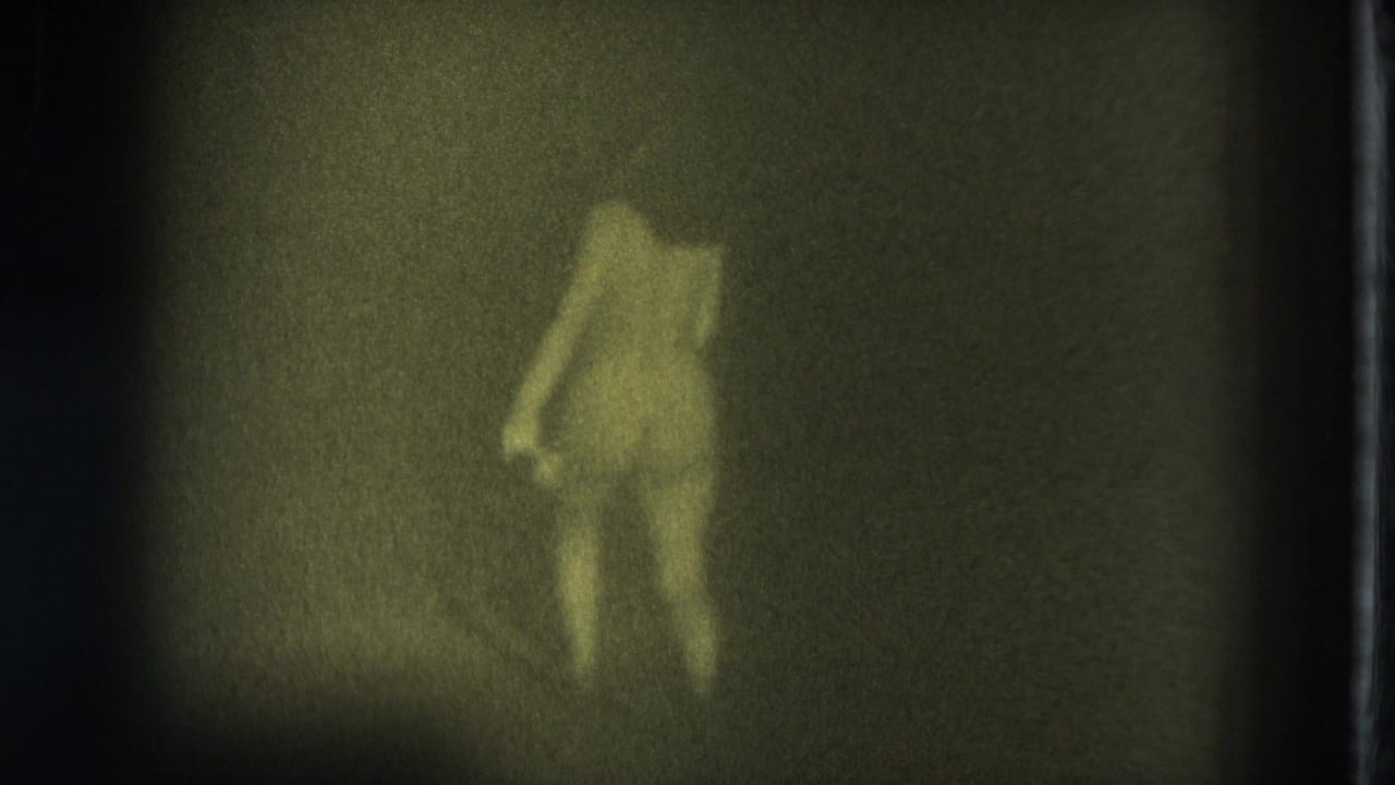 1975 San Francisco Super 8 frame with a nude adult woman walking across a dim stage, partly softened by grain and shadow.