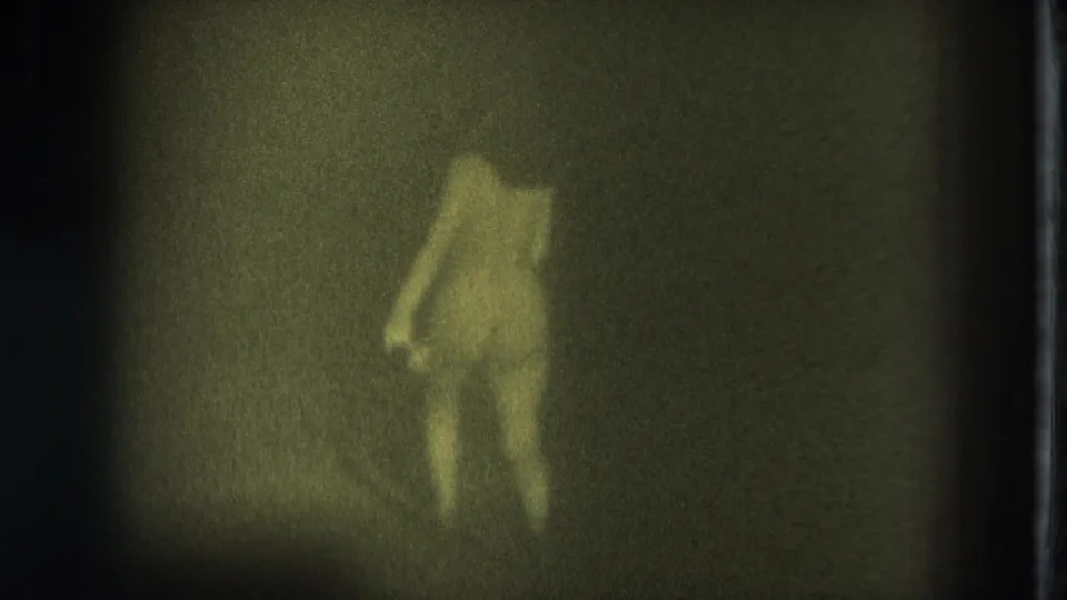 1975 San Francisco Super 8 frame with a nude adult woman walking across a dim stage, partly softened by grain and shadow.