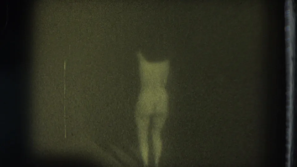 1975 San Francisco Super 8 frame showing the nude adult performer from behind in a dark, low-exposure stage setting.