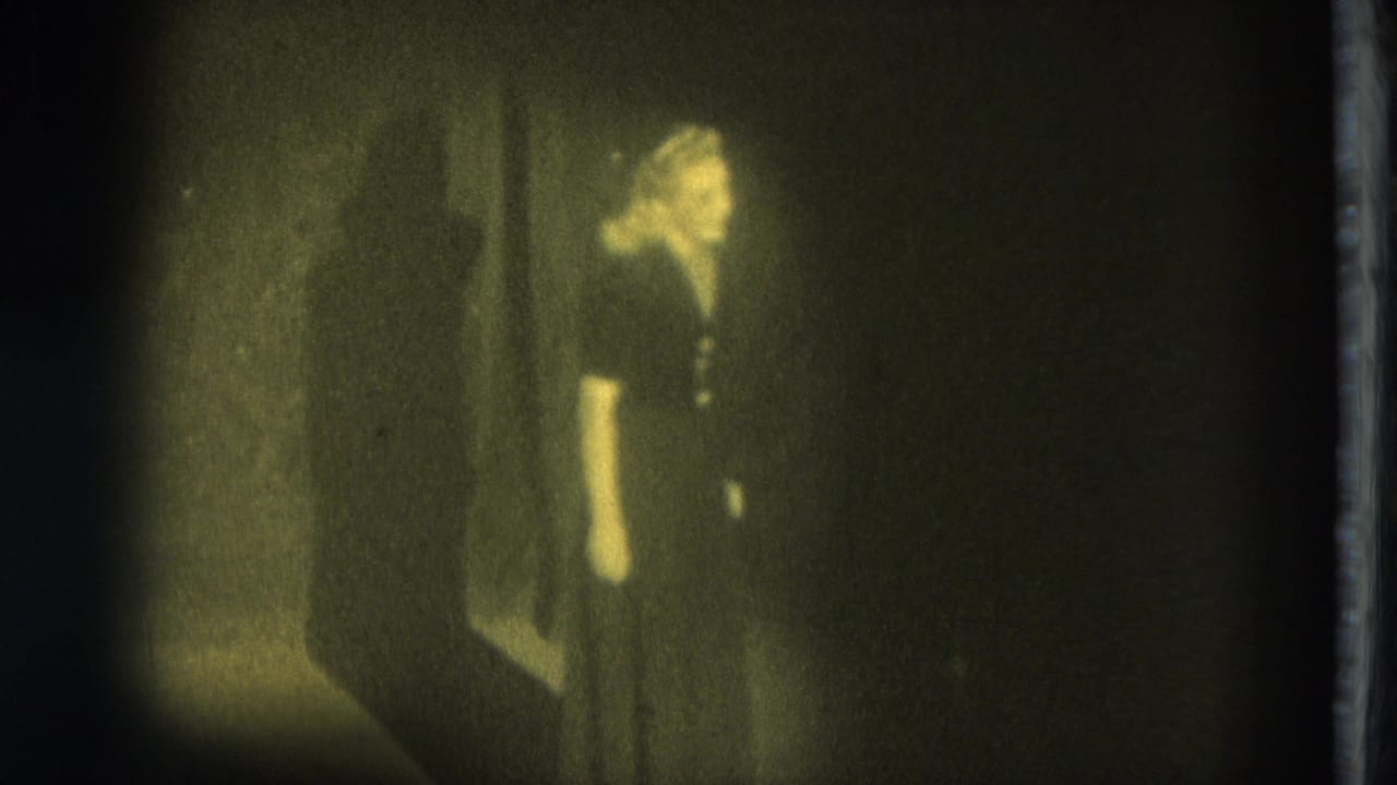 Adult woman in a dark dress stands in dim light beside a wall or curtain.