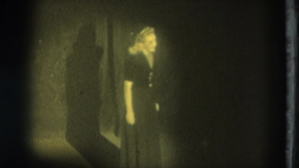 1975 San Francisco 8mm frame showing an adult woman performer in a dark dress under dim yellow light, with a shadowed wall beside her.