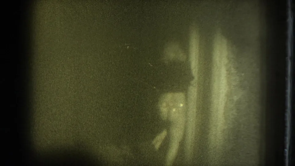 Grainy 1975 San Francisco 8mm frame showing the performer partly low in frame near vertical curtains or fabric, with details obscured by blur and exposure.