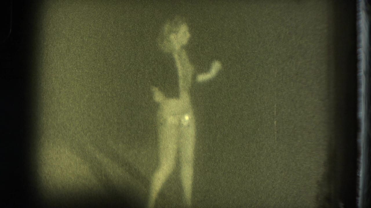 Female performer in light undergarments dances against a dark stage backdrop in grainy 8mm footage from San Francisco, 1975.