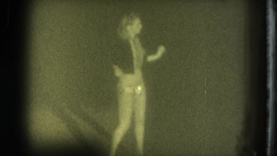 Female performer in light undergarments dances against a dark stage backdrop in grainy 8mm footage from San Francisco, 1975.