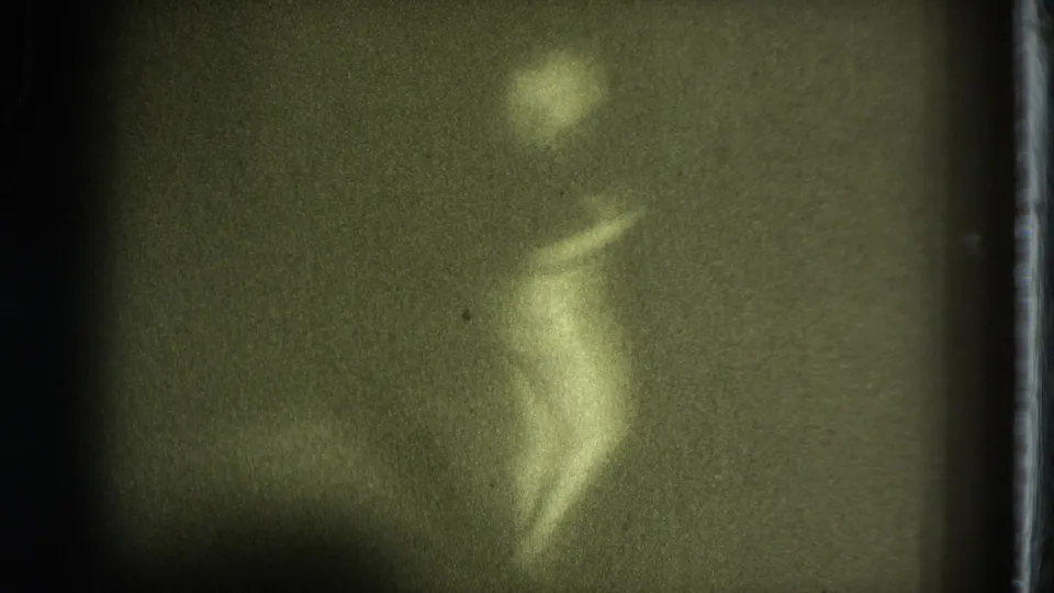 A blurred female performer moves through a dance pose on a low-lit stage, with heavy grain and green tint from the 8mm scan.