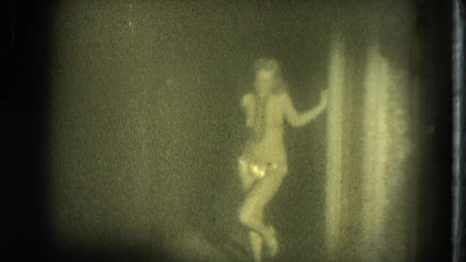 In 1975 San Francisco 8mm footage, a woman in a light outfit poses beside a curtain under soft greenish-yellow light.