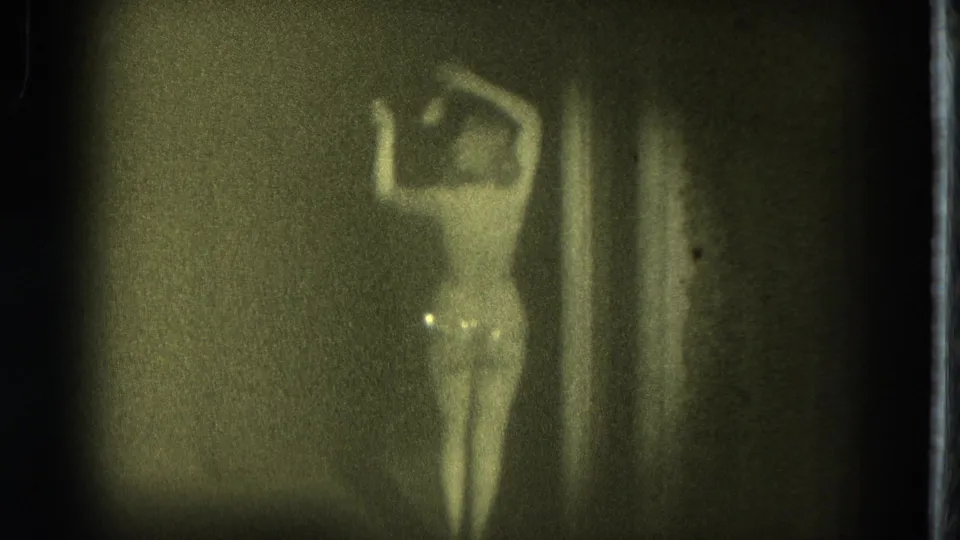 A woman in a light outfit lifts both arms near a curtain, captured in soft, grainy 8mm footage from San Francisco in 1975.