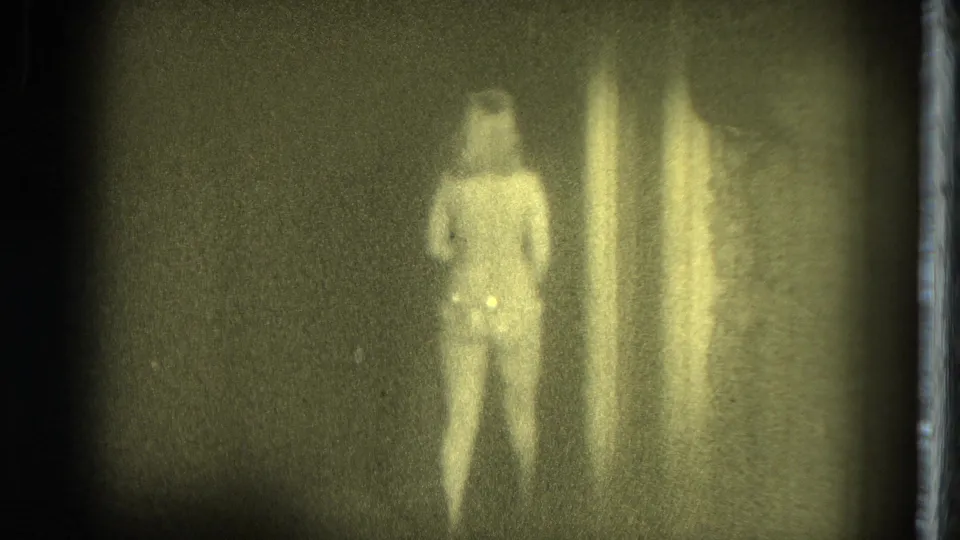 The performer stands with her back toward the camera beside a curtain, seen through soft, greenish 1975 8mm home-movie texture.