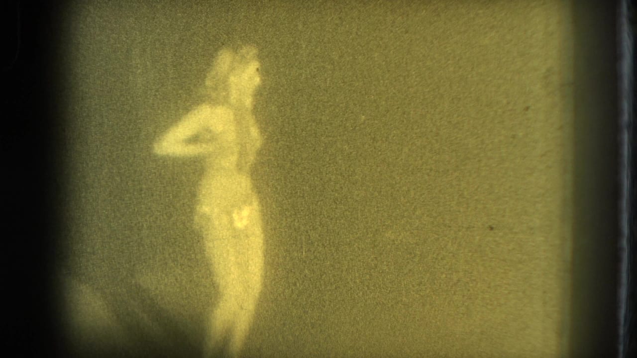 1975 San Francisco 8mm frame showing an adult woman in minimal clothing posing in profile under dim yellow-green exposure.