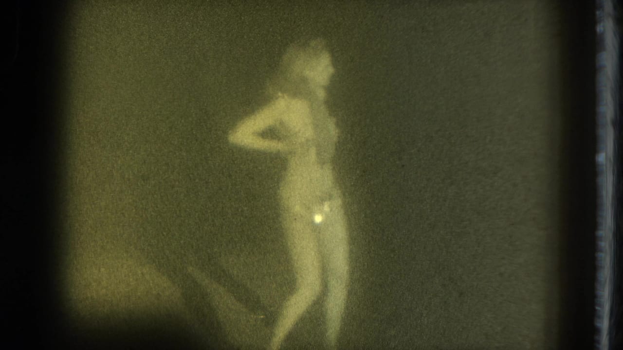 1975 San Francisco 8mm frame of an adult woman in minimal clothing standing in profile with one arm held back.