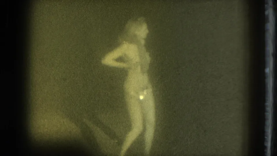 1975 San Francisco 8mm frame of an adult woman in minimal clothing standing in profile with one arm held back.