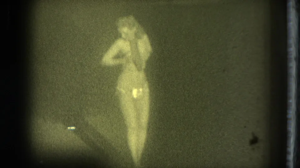 1975 San Francisco 8mm frame showing an adult woman turning slightly as she poses under dim, warm-tinted light.