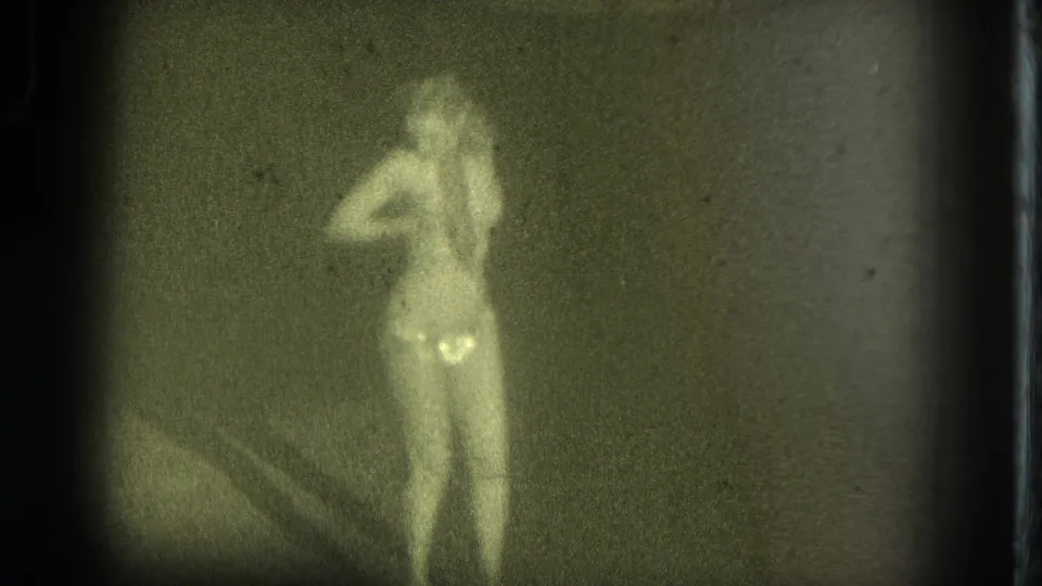 A female performer appears in soft focus in 1975 San Francisco footage, standing under dim light with bright costume highlights.