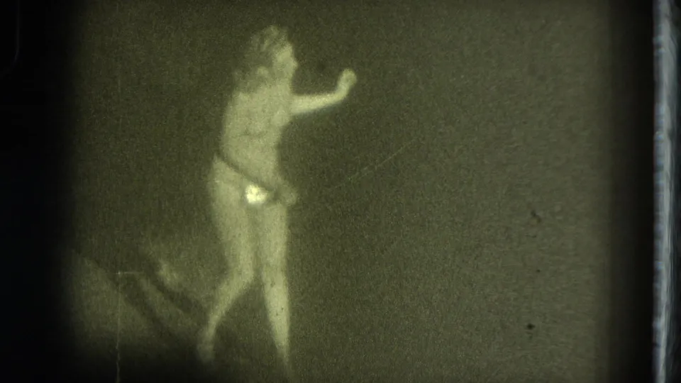 A dancer or entertainer appears in a dim 1975 San Francisco frame, with one arm extended and reflective costume details visible.