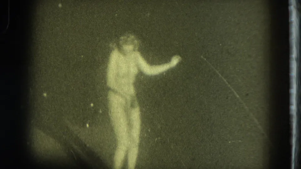 A female performer in a dim 1975 San Francisco frame stands in profile, with a bent arm and soft film-worn detail.