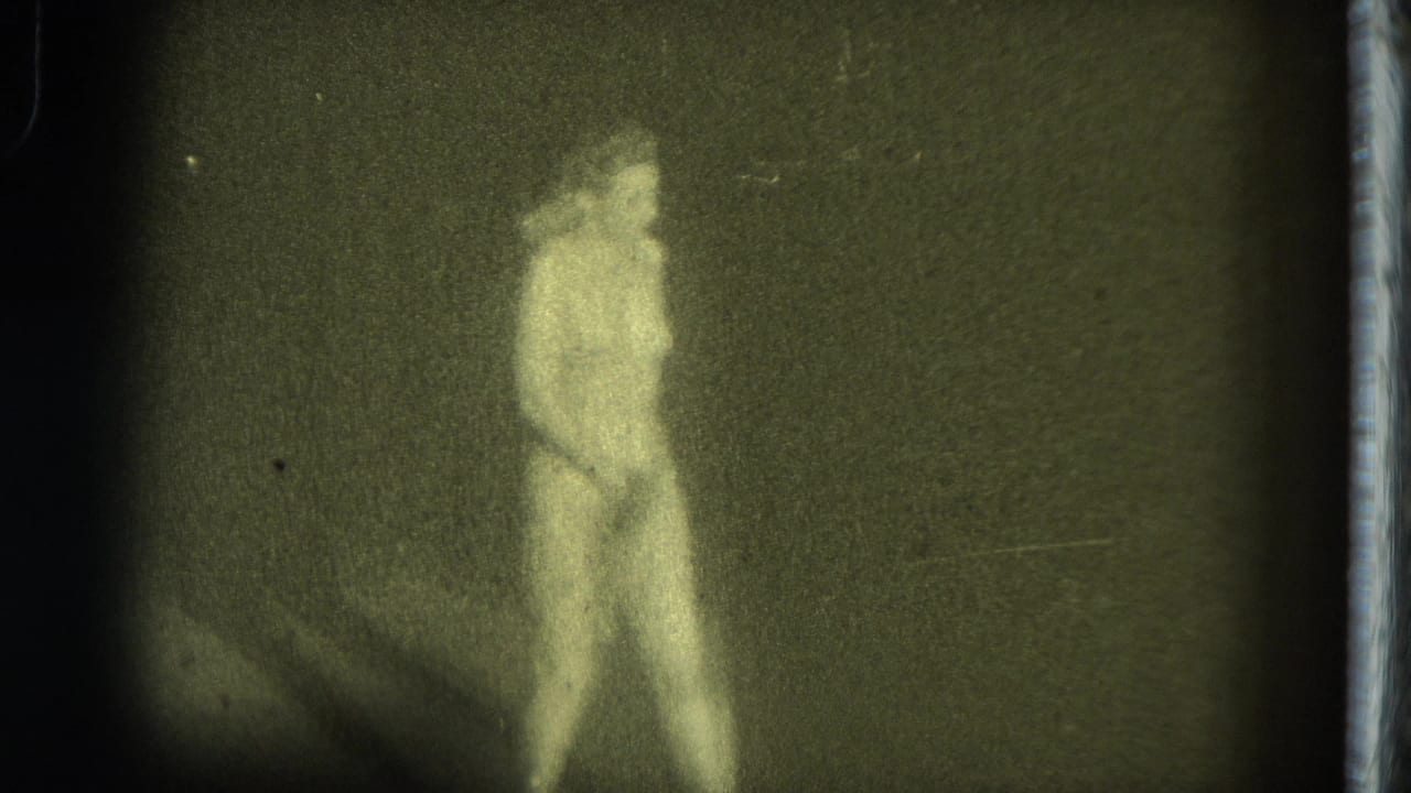 1975 8mm footage from San Francisco showing a nude burlesque performer standing on a dark stage in soft, grainy light.