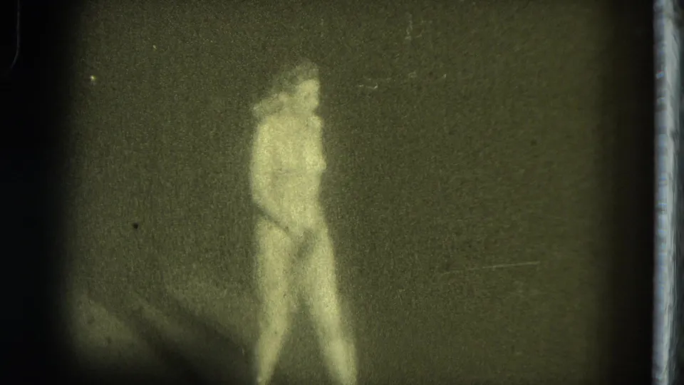 1975 8mm footage from San Francisco showing a nude burlesque performer standing on a dark stage in soft, grainy light.