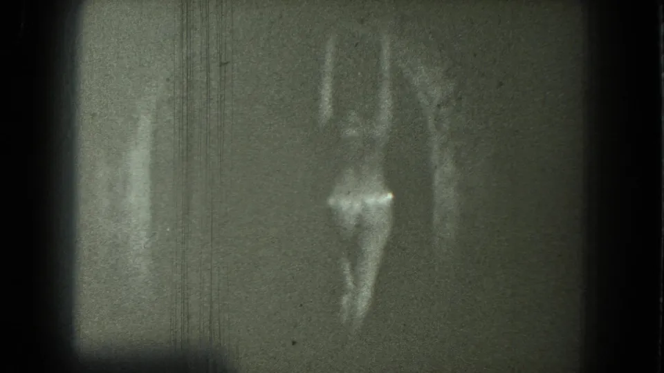 1975 San Francisco 8mm frame with an adult woman in shimmering revealing clothing dancing near a pale vertical form in soft focus.