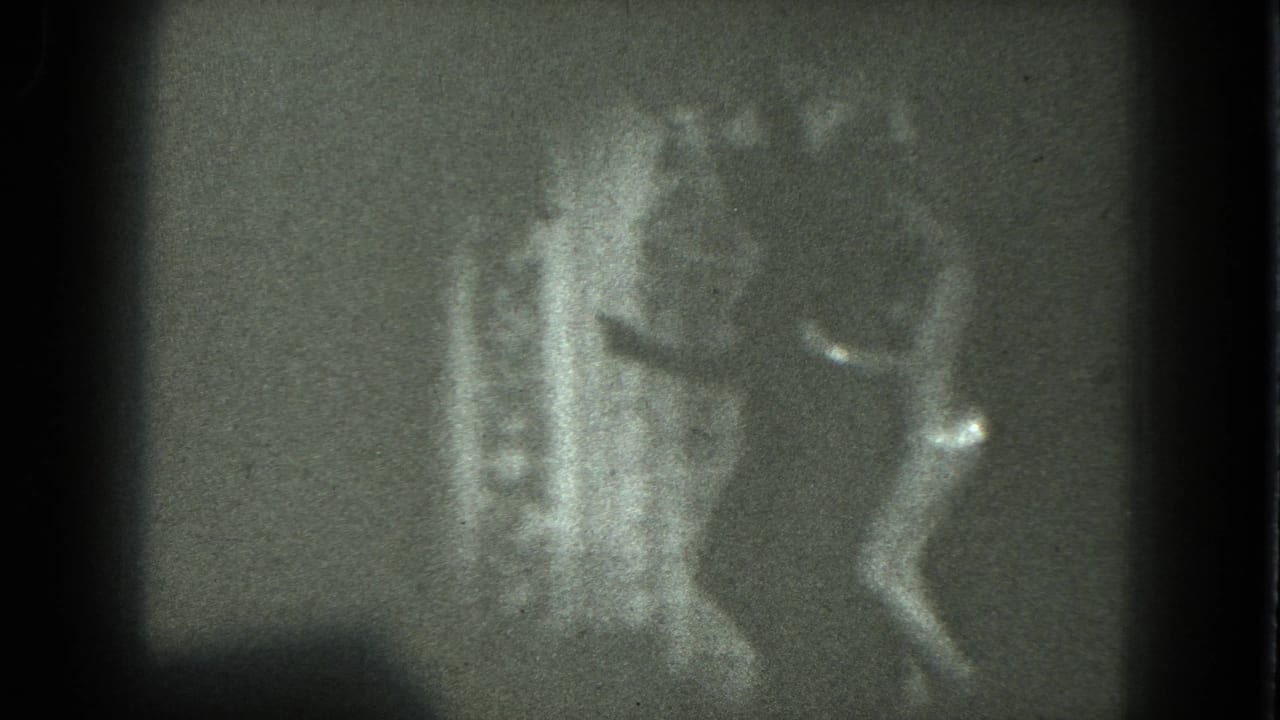 1975 San Francisco 8mm frame showing a nude adult woman in heels moving beside a plain backdrop under strong, soft-edged light.
