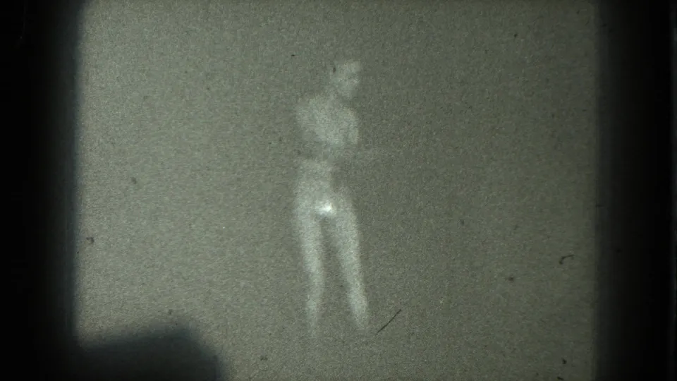 Soft, grainy 1975 San Francisco 8mm frame showing a nude adult woman in heels standing before a plain backdrop.