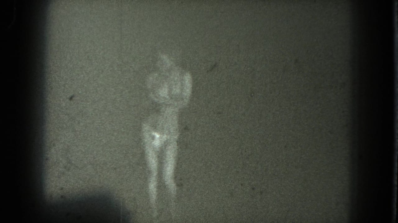 The faint figure of the dancer is visible in a low-light 8mm frame, with one arm raised and details obscured by grain.