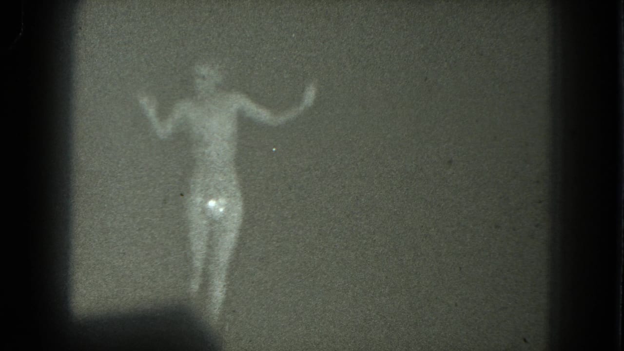 Soft 8mm frame of a nude adult dancer with both arms raised in a dim room, from metadata-linked 1975 San Francisco footage.
