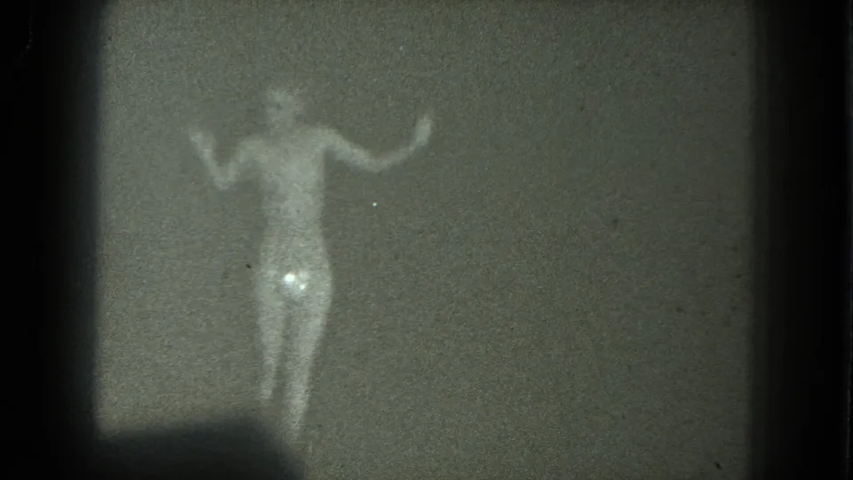 Soft 8mm frame of a nude adult dancer with both arms raised in a dim room, from metadata-linked 1975 San Francisco footage.