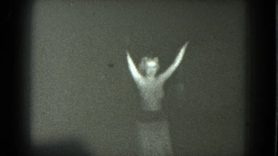 Adult woman performing on a dim San Francisco stage in 1975, seen through grainy 8mm footage with arms lifted.