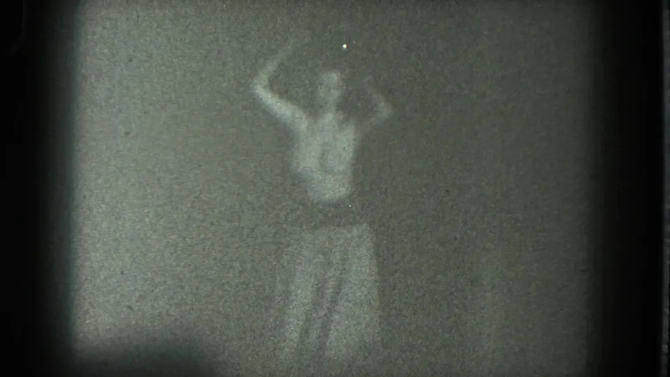 Adult performer in grainy 8mm footage gestures with one arm raised on a dim San Francisco stage.