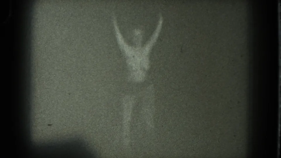 Adult woman steps back on a dim 1975 San Francisco stage, arms lifted as the grainy 8mm frame darkens at the edges.
