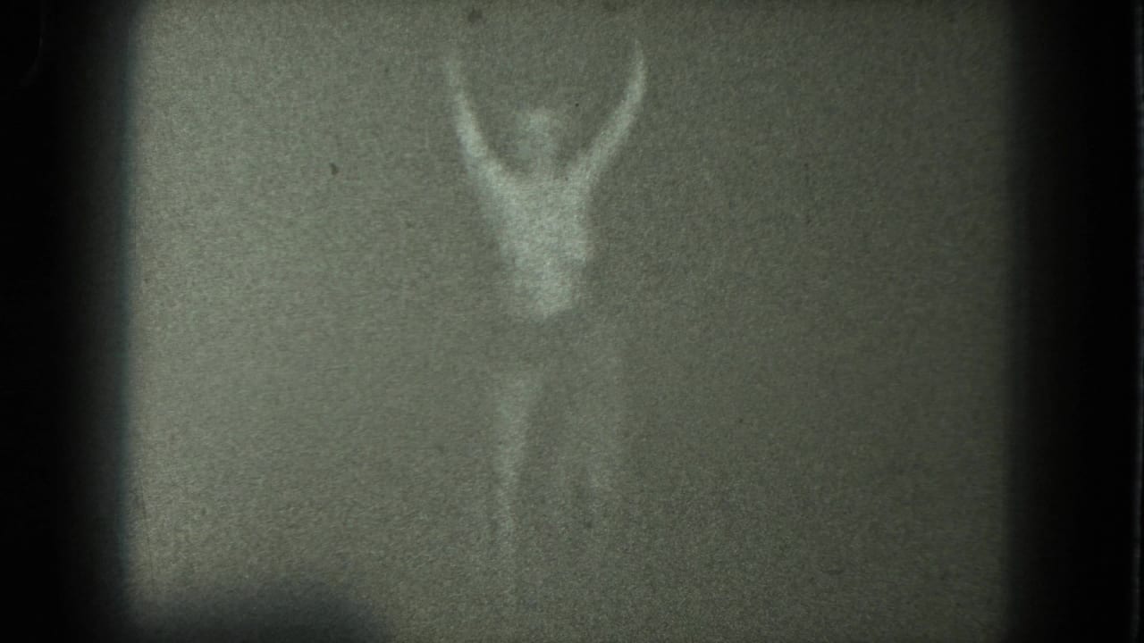 Dim nude female figure with arms raised against a pale curtain or wall.