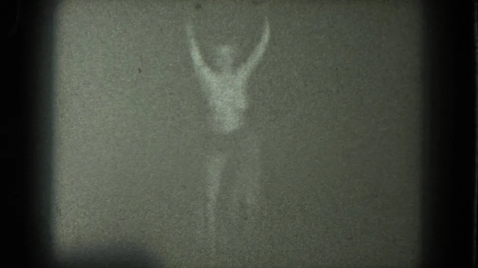 1975 San Francisco 8mm frame showing a soft, grainy adult female figure with raised arms in a dim interior.