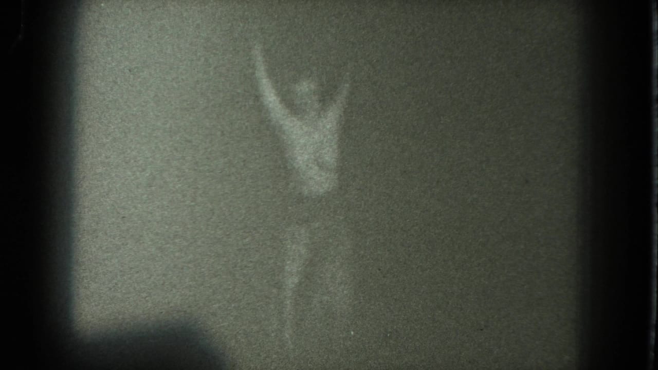 Grainy 1975 San Francisco 8mm frame with an adult figure holding raised arms in a low-light room.