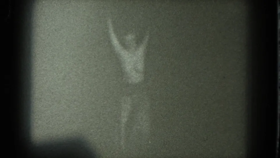Grainy 1975 San Francisco 8mm frame with an adult figure holding raised arms in a low-light room.