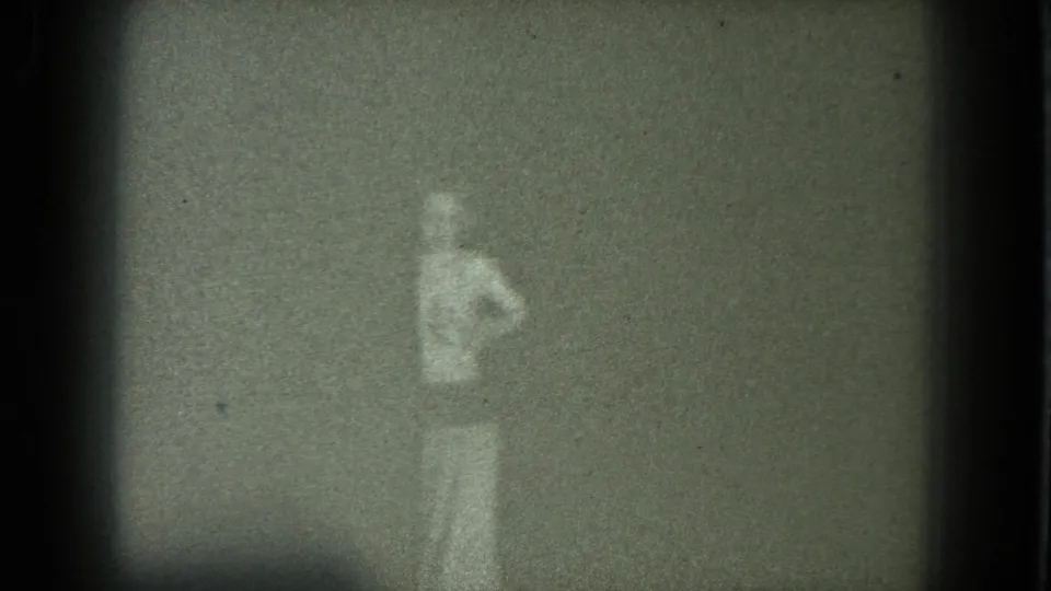 1975 San Francisco home-movie frame showing a blurred adult figure lowering their arms in a dark, grainy interior.