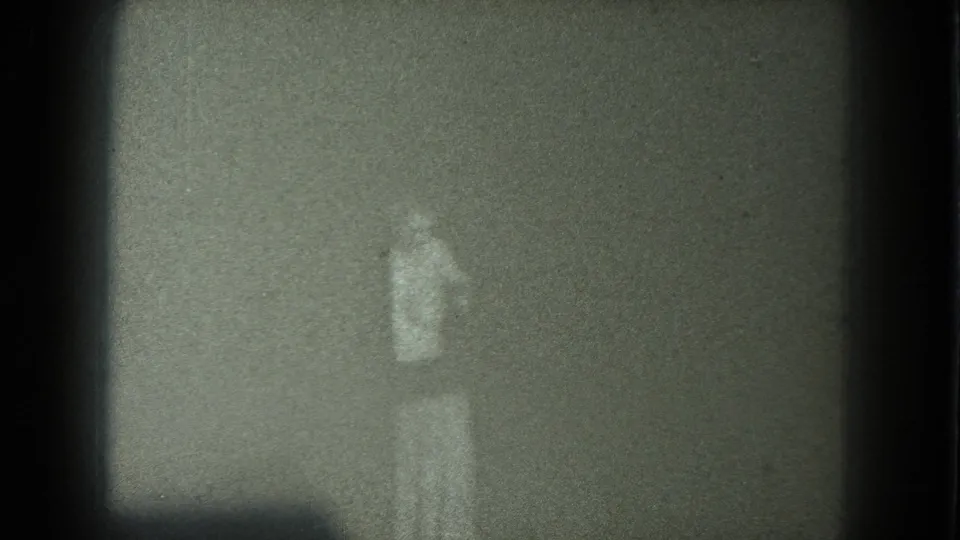Faint standing person in light clothing within a dim 1975 8mm San Francisco home-movie frame.