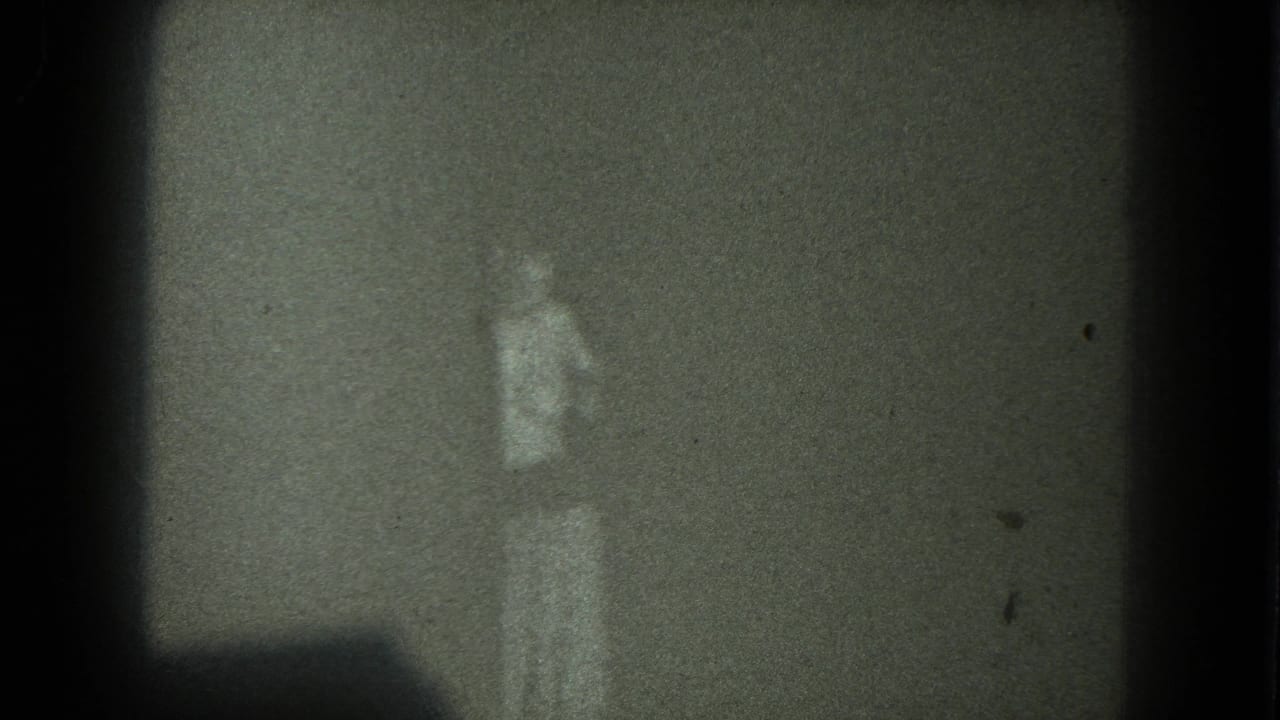 Low-light 8mm frame showing a faint person in light clothing near a shadowed corner, San Francisco metadata supported.