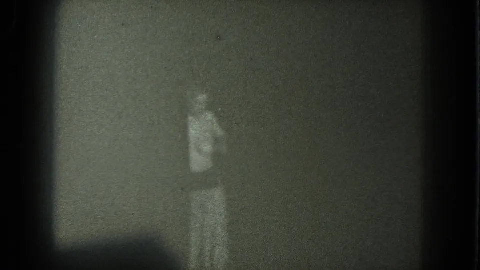 Grainy 1975 8mm home-movie frame with an indistinct standing person in light clothing against a dark background.