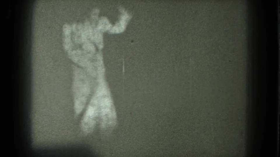Adult woman bends forward during a dance-like movement in grainy 8mm home-movie footage, with partial undress visible.